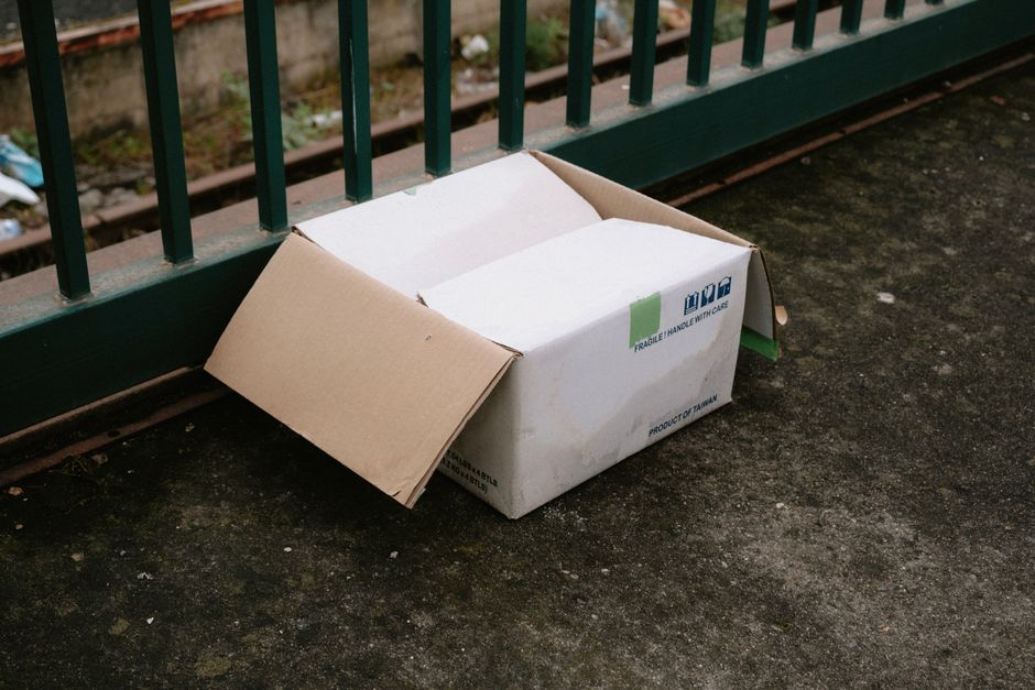 A white cardboard box with partially open flaps, containing smaller pieces of cardboard, is placed on a dark, weathered pavement surface next to a green metal railing. The box has printed labels indicating it is a product of Taiwan and features a green fragile handling sticker. The surrounding area appears to be outdoors, possibly on a bridge or platform, with a blurred view of railway tracks and gravel in the background. The scene suggests a scenario of private waste disposal or on-site rubbish collection, with Waste Clearance Barnes often handling such bulky and cardboard waste outside typical collection points, supporting alternative rubbish removal methods in the context of Barnes Bridge station.