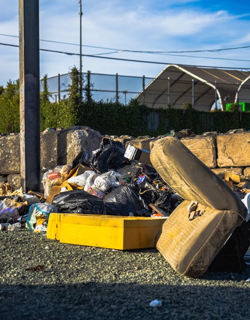 A collection of discarded rubbish is piled on a paved outdoor surface next to a low stone wall. The debris includes black plastic garbage bags, a yellow plastic crate, a worn-out car seat with fabric upholstery, and various smaller waste items such as packaging and paper. The pile appears to be an informal or private waste collection site, with the items loosely arranged and spilling onto the ground. In the background, there is a metal utility pole with power lines extending across the scene, and a mesh fence partially obscured by greenery. Behind the fence, a large, curved, semi-transparent structure is visible, suggesting an industrial or recreational area. The scene is illuminated by natural daylight, with a blue sky and some wispy clouds overhead. This image exemplifies an on-site waste clearance situation where independent rubbish disposal methods are used, aligning with services offered by companies like Waste Clearance Barnes to manage bulky waste removal professionally and efficiently.
