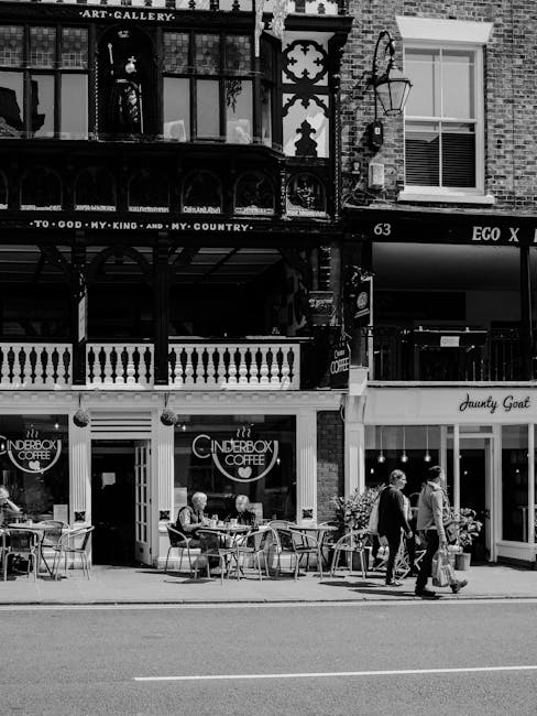 A black and white photograph depicts a street-level commercial building with multiple retail and hospitality establishments. In the foreground, there is an outdoor seating area belonging to The Tinderbox Coffee, featuring several metal chairs and round tables arranged on the sidewalk. Several patrons are seated, and a couple of pedestrians are walking past, one carrying a shopping bag. The building has a brick facade with large display windows and signs for different businesses, including The Tinderbox Coffee and Dainty Goat. Above, there are additional floors with balconies, decorative ironwork, and windows, some with blinds drawn. A vintage-style street lamp is attached to the building, and there is a signage for Eco X. The scene appears urban, with a mix of commercial activity and pedestrian movement typical of a busy shop and cafe area. The image conveys a snapshot of street-level cafe culture and the surrounding architecture, which may be relevant to private waste management or rubbish removal services for such busy retail and hospitality spaces managed by companies like Waste Clearance Barnes.