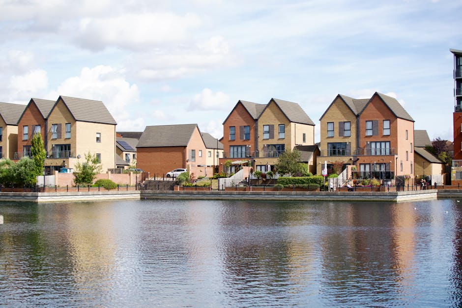 A row of modern, multi-storey residential houses with pitched roofs, constructed from brick in shades of beige, red, and light brown, line the edge of a calm river. The houses are positioned on a paved walkway or terrace, with small landscaped gardens and outdoor steps leading down to the water. One house features a glass balcony on the upper floor, while another has a small front garden enclosed with a low brick wall. The river reflects the buildings and the partly cloudy sky above, with soft light illuminating the scene. The environment appears calm and orderly, showcasing a picturesque riverside setting. To the far right, part of a taller apartment building is visible, highlighting the urban context. This scene exemplifies private residential properties that may require external rubbish removal or clearance services, contributing to maintaining the neighborhood’s tidy appearance in line with waste clearance operations by Waste Clearance Barnes.